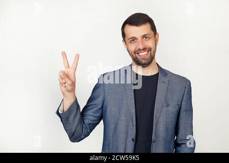 Young businessman man with a beard in a jacket, smiling, looking at the camera, showing thumbs up, making a victory sign. Number two. Portrait of a man on a gray background. Stock Photo
