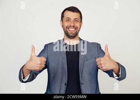 Young businessman man with a beard in a jacket, a sign of success makes a positive gesture with his hand, thumbs up smiling and happy. Cheerful expression and winner gesture. gray background Stock Photo