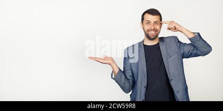 Young businessman man with beard smiling and happy with open palm showing copy space and pointing finger to forehead. I'm thinking about it. Portrait of a man on a gray background. Stock Photo