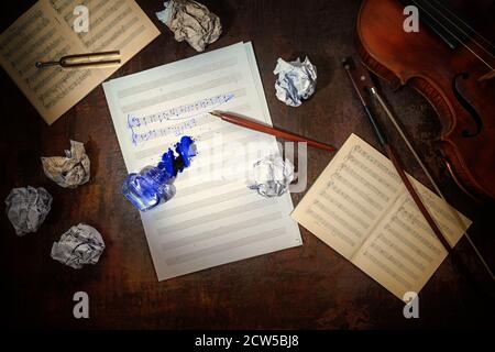 Violin, plain and crumpled sheet of music, handwritten beginning of a musical composition and a overturned inkwell on a dark brown table, workplace of Stock Photo
