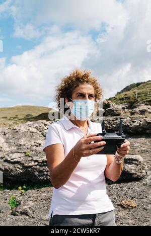 Middle aged woman wearing face mask navigating a flying drone with remote control. Woman in nature flying drone and taking pictures. Stock Photo