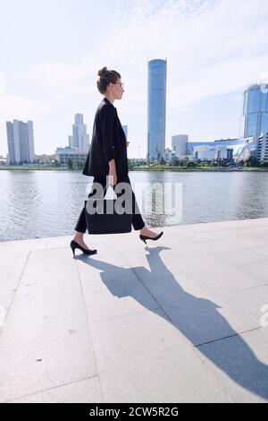 Young elegant businesswoman in formalwear moving along riverside on sunny day Stock Photo