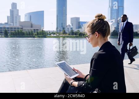 Young contemporary businesswoman using tablet while sitting by riverside Stock Photo