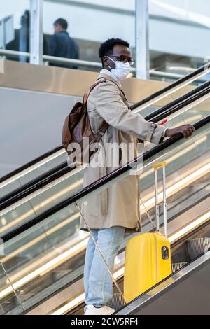 Afro-American traveler man with yellow suitcase stands on escalator in airport terminal, wear face medical mask to protect yourself from contact with Stock Photo
