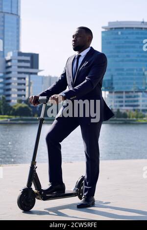 Young businessman in formalwear standing on electric scooter by riverside Stock Photo