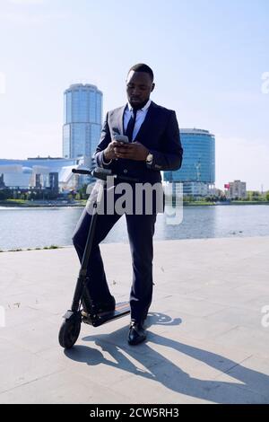 Young elegant businessman of African ethnicity standing on electric scooter