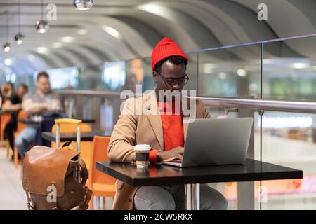 Stylish African American traveler millennial man in beige coat and red hat resting, sitting at cafe table in airport terminal, works remotely on lapto Stock Photo