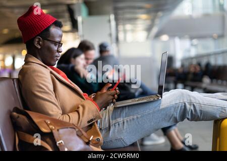 Side view of stylish African American traveler millennial man resting and sitting in hall of airport, using phone, works remotely on laptop while wait Stock Photo