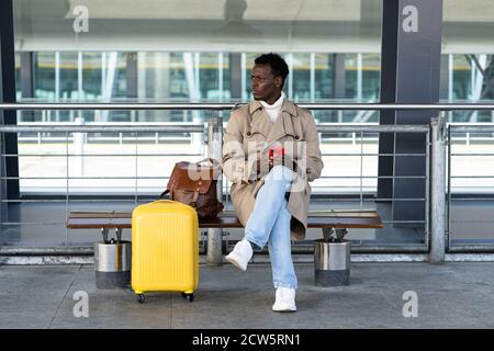 Afro-American traveler man with suitcase sitting on bench in airport terminal or railway station, using mobile phone, calling taxi, waiting public tra Stock Photo