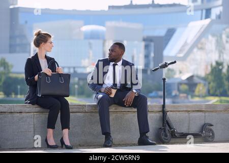 Two young intercultural business partners in formalwear sitting by riverside Stock Photo
