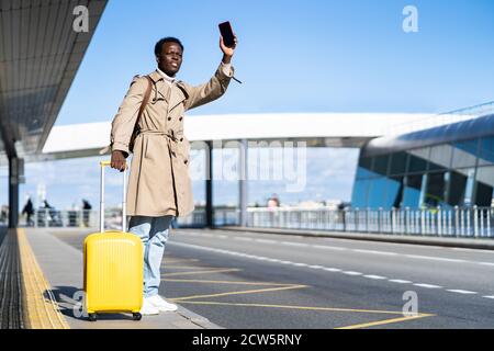 Afro-American millennial traveler man wear beige trench coat with yellow suitcase stands in airport terminal, holding phone and calling taxi, raising Stock Photo