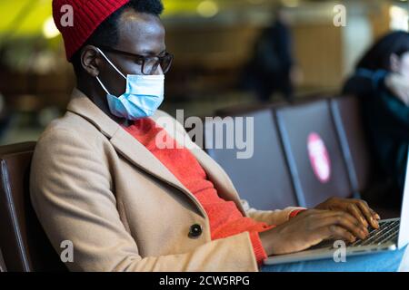 Stylish Black traveler millennial man wear face protective mask, sitting in hall of airport, works remotely on laptop while waiting for a flight and b Stock Photo