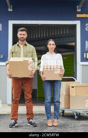 Vertical full length portrait of young couple holding boxes and looking at camera while standing by self storage facility Stock Photo