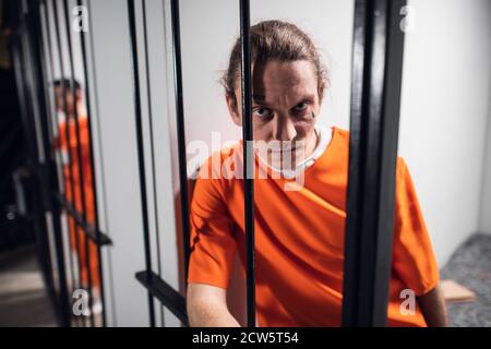 A desperate criminal maniac with tattoos in a solitary cell is kept behind bars. A hard look at the camera Stock Photo