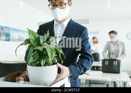 Young Asian businessman in formalwear and protective mask taking out flowerpot Stock Photo