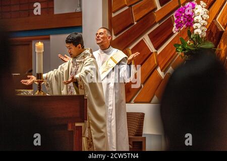 Wearing ceremonial robes, a deacon assists an Asian American priest conducting mass at the altar of a Southern California Catholic church. Stock Photo