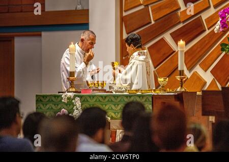 Wearing ceremonial robes, a deacon assists an Asian American priest preparing for communion during mass at the altar of a Southern California Catholic Stock Photo