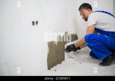 The worker attaches the tiles to the wall with cement. He is wearing a blue work clothers and black gloves Stock Photo