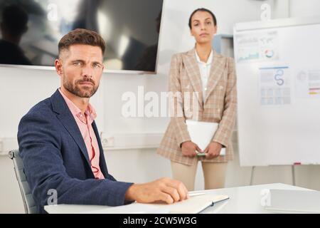Portrait of handsome businessman looking at camera while sitting at table in conference room with female colleague or secretary in background, copy space Stock Photo