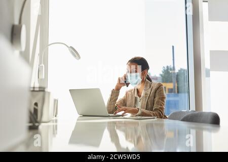 Portrait of mixed-race businesswoman wearing mask and speaking by smartphone while working in white office, copy space Stock Photo