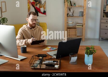 Young bearded creative freelance designer sitting by table in front of laptop Stock Photo