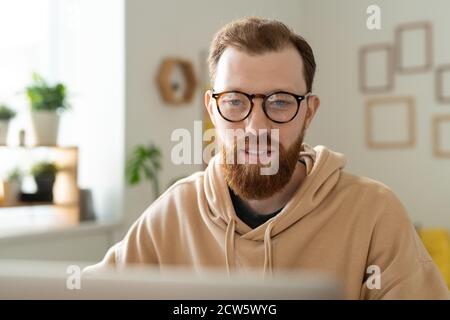 Young bearded freelance designer in eyeglasses looking at laptop display Stock Photo