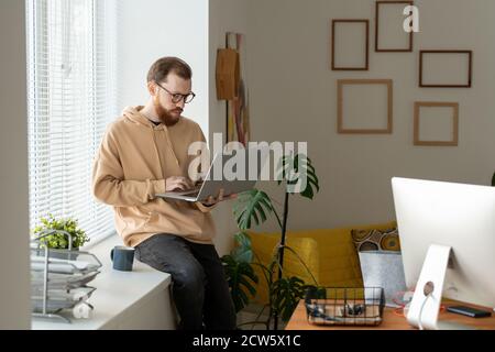 Young bearded freelancer using laptop while networking on windowsill Stock Photo