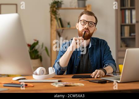 Young bearded pensive freelancer with stylus by his mouth working at home Stock Photo