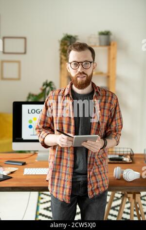 Young bearded creative designer using tablet and stylus against computer monitor Stock Photo