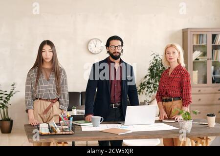 Three intercultural colleagues in smart casualwear standing in row along table Stock Photo