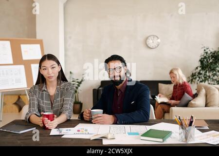 Two young elegant colleagues with mugs of coffee or tea sitting by workplace Stock Photo