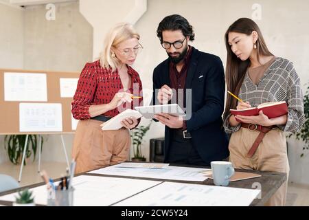 Two businesswomen making notes and looking at display of tablet held by man Stock Photo