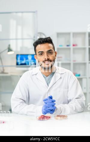 Young gloved male scientific researcher in whitecoat looking at you by workplace Stock Photo