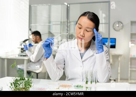 Young tired Asian female researcher in whitecoat and gloves touching her head Stock Photo