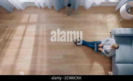 Young Man is Sitting on a Floor, Working or Studying on a Laptop. Cozy Living Room with Modern Interior, Grey Sofa and Wooden Flooring. Top View Stock Photo
