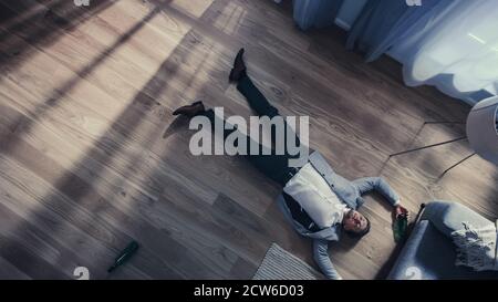 Poor Depressed Drunk Young Man is Crawling Towards a Sofa in an Apartment with Wooden Flooring. He's Holding a Bottle of Beer and Tries to Drink the Stock Photo