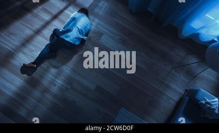 Poor Depressed Drunk Young Man is Sleaping on the Floor Near Sofa in an Apartment with Wooden Flooring. Dramatic Top View Camera Shot. Stock Photo