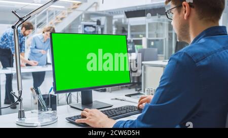 Over the Shoulder Shot of Engineer Working with Green Mock-up Screen Desktop Computer. In the Background Engineering Facility with Specialists Working Stock Photo