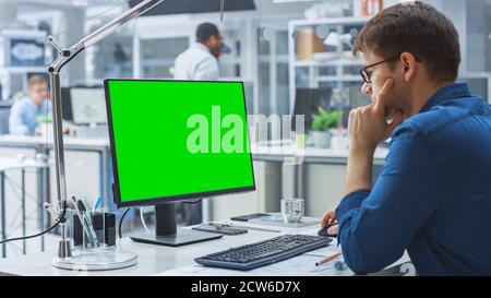 Over the Shoulder Shot of Engineer Working with Green Mock-up Screen Desktop Computer. In the Background Engineering Facility with Specialists Working Stock Photo