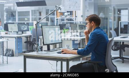 Over the Shoulder Shot of Engineer Working with CAD Software on Desktop Computer, Screen Shows Technical Drafts and Drawings. In the Background Stock Photo