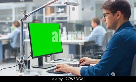 Over the Shoulder Shot of Engineer Working with Green Mock-up Screen Desktop Computer. In the Background Engineering Facility with Specialists Working Stock Photo