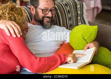 Adult people caucasian man and woman couple at home reading a book together sharing time and life on the couch - happy relationship lifestyle concept