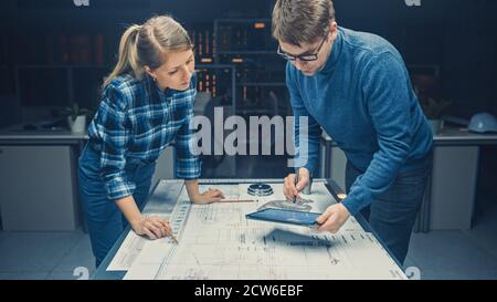 In the Dark Industrial Design Engineering Facility Male and Female Engineers Talk and Work on a Blueprints Using Conference Table. On the Desktop Stock Photo