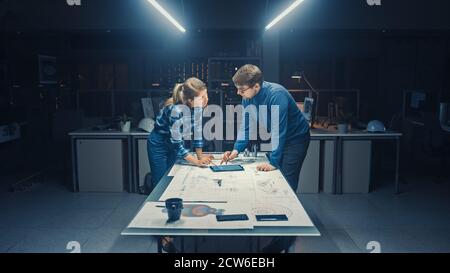 In the Dark Industrial Design Engineering Facility Male and Female Engineers Talk and Work on a Blueprints Using Conference Table. On the Desktop Stock Photo