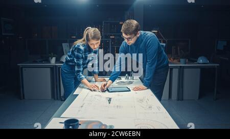 In the Dark Industrial Design Engineering Facility Male and Female Engineers Talk and Work on a Blueprints Using Conference Table. On the Desktop Stock Photo
