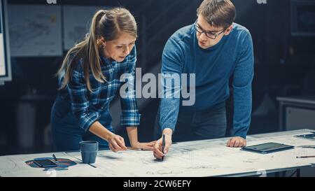 In the Dark Industrial Design Engineering Facility Male and Female Engineers Talk and Work on a Blueprints Using Digital Tablet and Conference Table Stock Photo