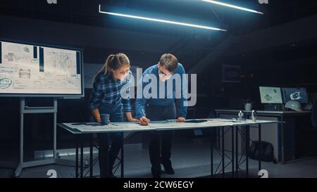 In the Dark Industrial Design Engineering Facility Male and Female Engineers Talk and Work on a Blueprints Using Digital Tablet and Conference Table Stock Photo
