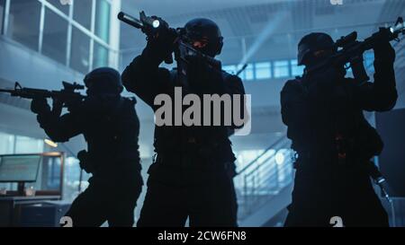 Masked Squad of Armed SWAT Police Officers Stand in Dark Seized Office Building with Desks and Computers. Soldiers with Rifles and Flashlights Surveil Stock Photo