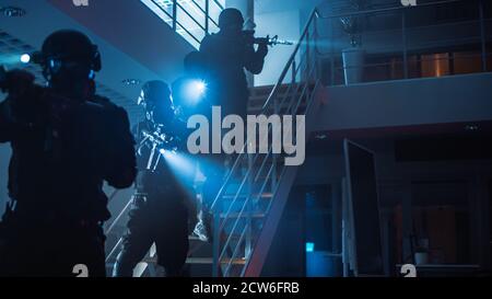 Masked Squad of Armed SWAT Police Officers Run Down the Stairs from a Second Floor in a Dark Office Building. Soldiers with Rifles and Flashlights Stock Photo