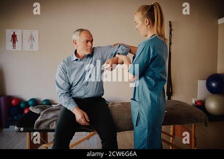 Young female physiotherapist helping senior man with elbow exercise in clinic - young female doctor helping senior patient Stock Photo